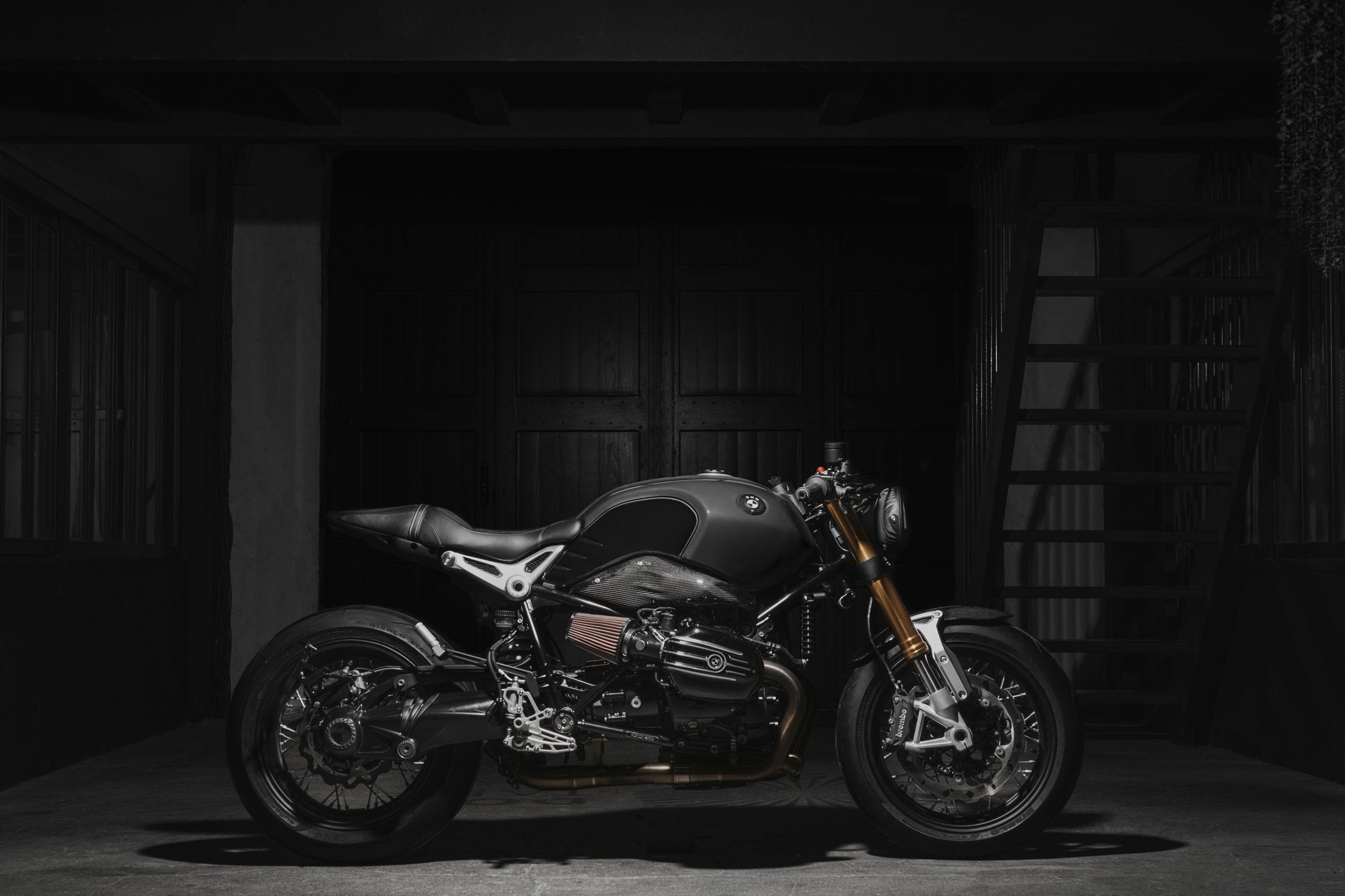BMW R nineT « 7075 Koffee » de profil dans un hangar sombre, tés de fourche usinés, boucle arrière aluminium et selle cuir 